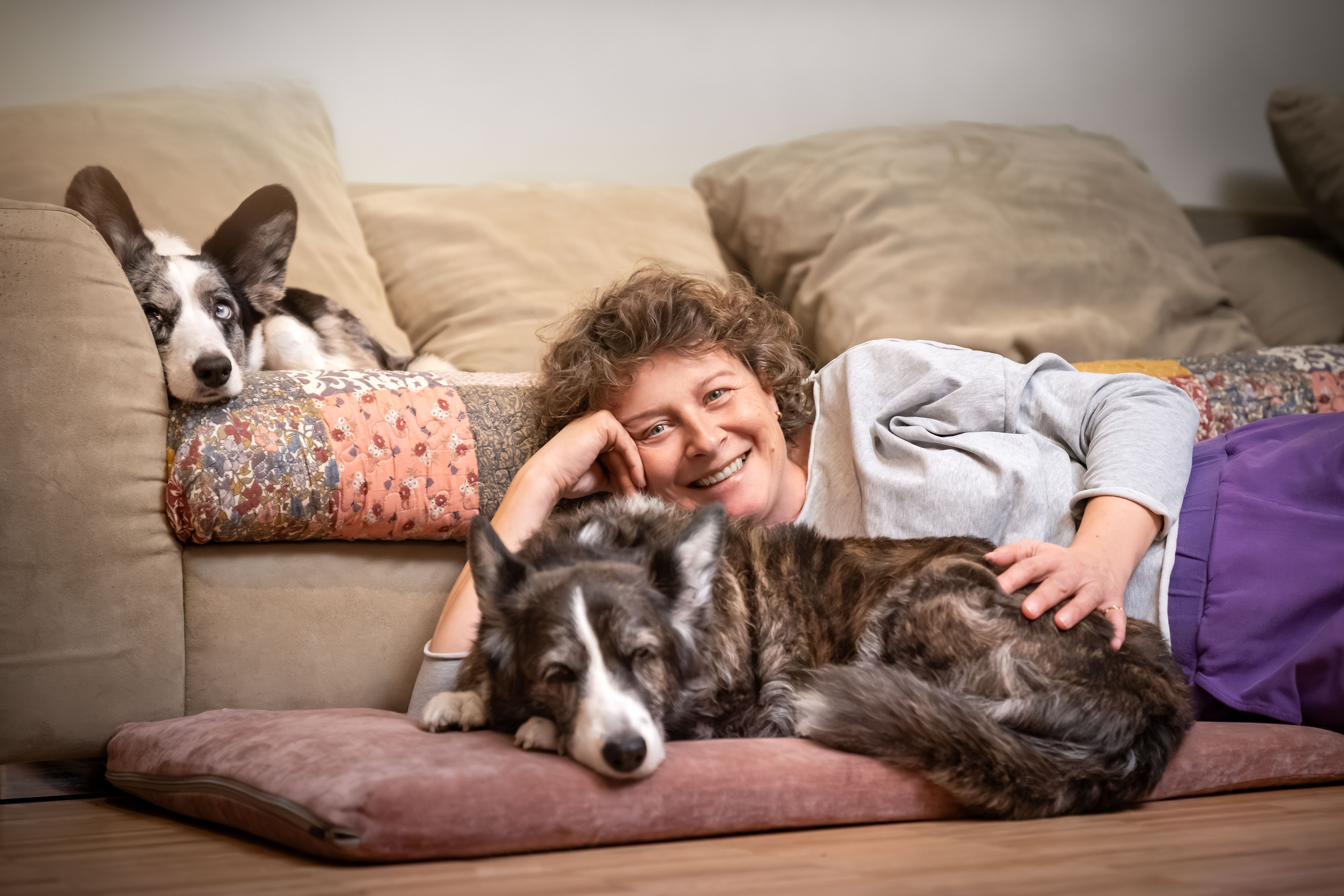 Petsitter z Warszawy uśmiecha się, spoczywając na kanapie z psem rasy welsh corgi cardigan, który leży obok niej na poduszce i patrzy w kamerę.